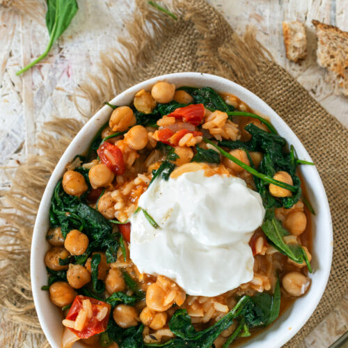 A white bowl filled with Mediterranean chickpea stew, featuring chickpeas, wilted spinach, diced tomatoes, and rice in a tomato-based sauce. The dish is topped with a generous dollop of creamy yogurt and set on a rustic background with scattered bread pieces and fresh spinach leaves.