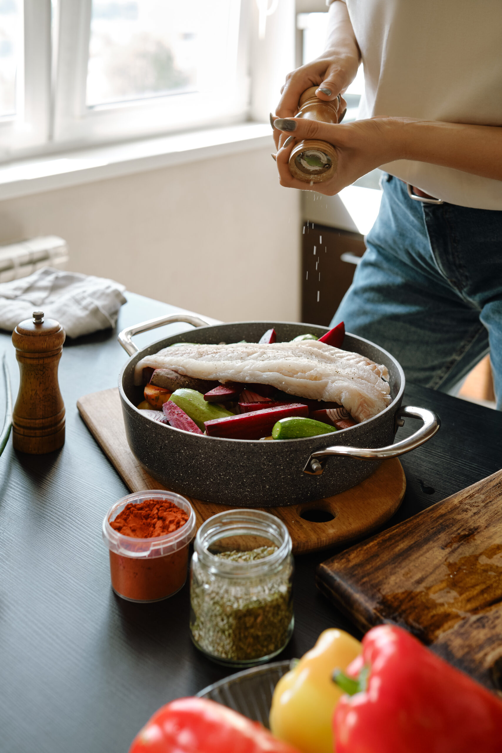 Hands adding pepper from hand spice mill to white fish fillet laying on top of vegetables in a frying pan